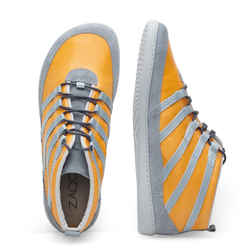 Ausgestellt sind zwei High-Top-Sneaker aus der ZAQQ-Kollektion. Einer ist von oben und der andere von der Seite zu sehen und gehört zur SPARQ Mid Yellow-Linie. Diese Sneaker aus Nappaleder haben einen leuchtend orangefarbenen Korpus, der durch hellgraue Akzente und Streifen sowie graue Sohlen und Schnürsenkel ergänzt wird. Im Inneren ist ein ZAQQ-Label zu sehen, das das unverwechselbare Barfußschuh-Design hervorhebt.