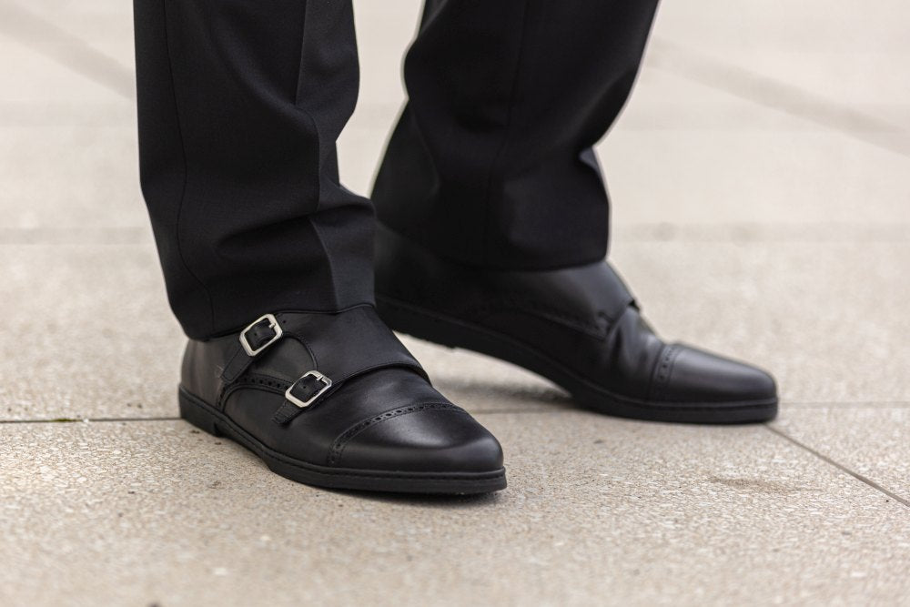 Eine Person in schwarzer Anzughose und eleganten ZAQQ MONQ Captoe Black-Schuhen mit stilvollen silbernen Schnallen steht auf einem gefliesten Bürgersteig. Der Fokus liegt auf den Schuhen, die ihr poliertes Aussehen und ihr elegantes Design hervorheben.