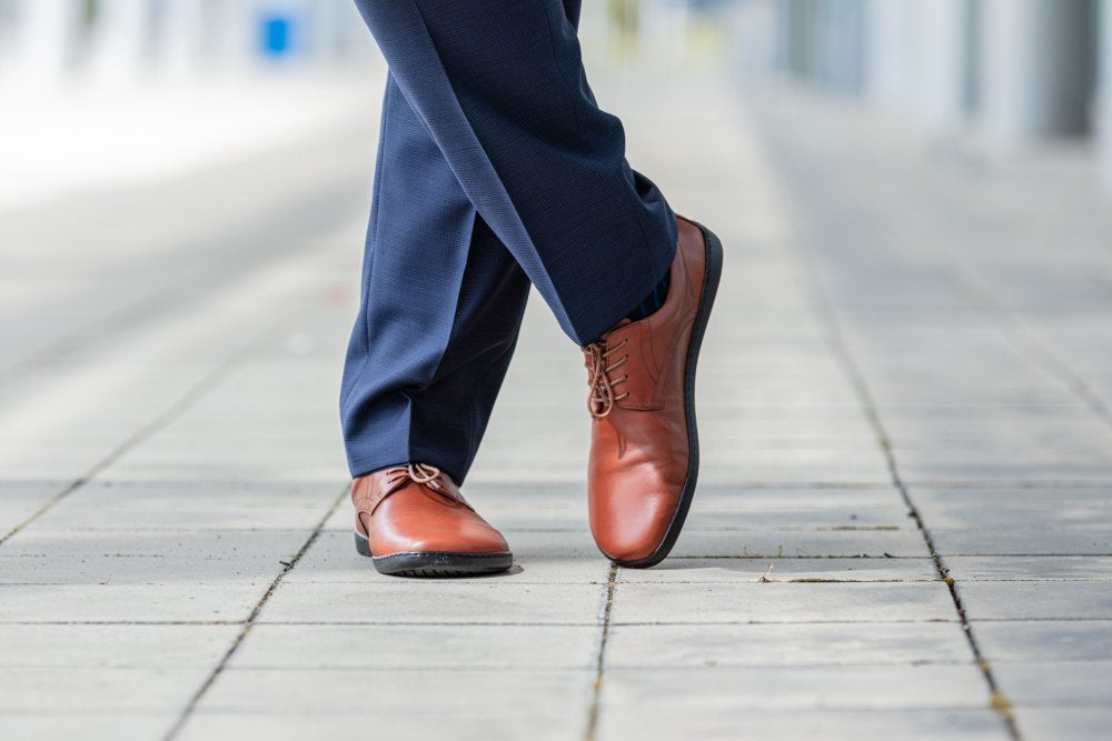 Nahaufnahme einer Person, die ZAQQs PEAQ Cognac-Lederschuhe und eine marineblaue Hose trägt und auf einem gepflasterten Gehweg spazieren geht. Das Bild fängt einen dynamischen Schritt ein und unterstreicht den Stil und die Eleganz dieser glatten Glattlederschuhe.