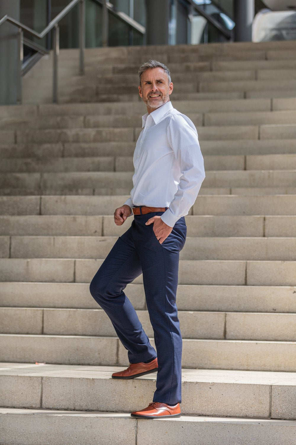 Ein Mann in weißem Hemd und marineblauer Hose steht selbstbewusst auf einer Treppe. Er trägt ZAQQs PEAQ Cognac-Schuhe und einen passenden braunen Gürtel. Mit der Hand in der Tasche zeigt der Hintergrund ein modernes Gebäude mit großen Fenstern.