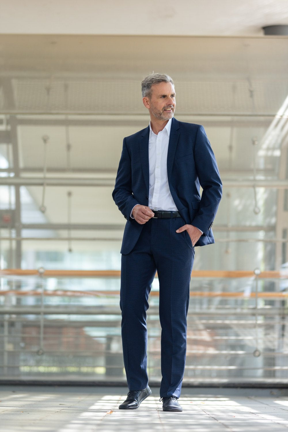 Ein Mann in blauem Anzug und weißem Hemd steht selbstbewusst da, die Hand in der Tasche, und blickt zur Seite. Seine schwarzen SEQUENT-Schuhe von ZAQQ glänzen im Licht und ergänzen die moderne Glasarchitektur im Hintergrund perfekt.