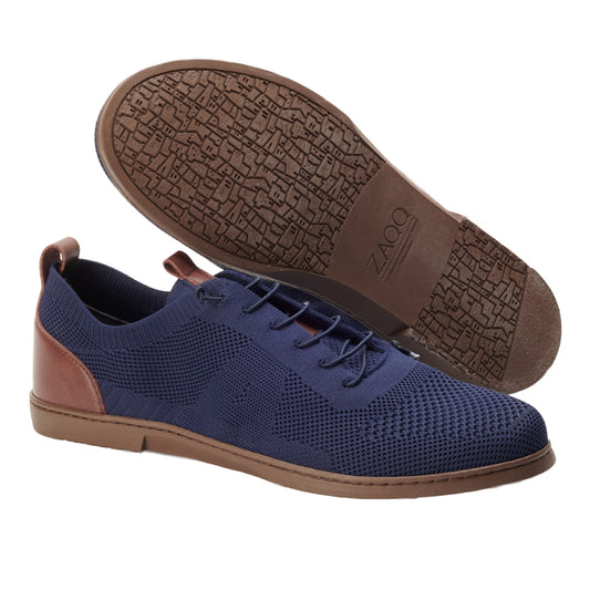 Ein Paar stylischer Herrenschuhe von ZAQQ im Design REQY Dark Blue, das aus einem blauen Strickstoff mit braunen Lederakzenten und innovativer Elastikschnürung besteht. Ein Schuh steht aufrecht und hebt sein Seitenprofil zusammen mit der strukturierten Sohle und dem Markenlogo hervor.