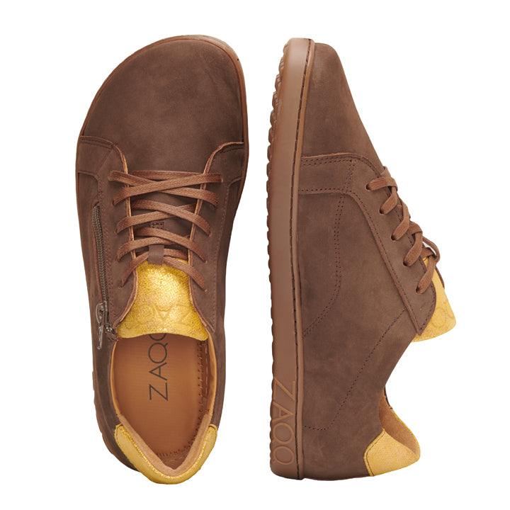 Ein Paar QOMFY Wide Brown-Sneaker, die auf Komfort ausgelegt sind, werden von der Seite und von oben präsentiert. Diese extra breiten Schuhe in Braun mit gelben Akzenten haben Schnürsenkel, einen seitlichen Reißverschluss und eine strukturierte Sohle und Spitze. Der Markenname ZAQQ ist auf der Innenseite eines Schuhs sichtbar.