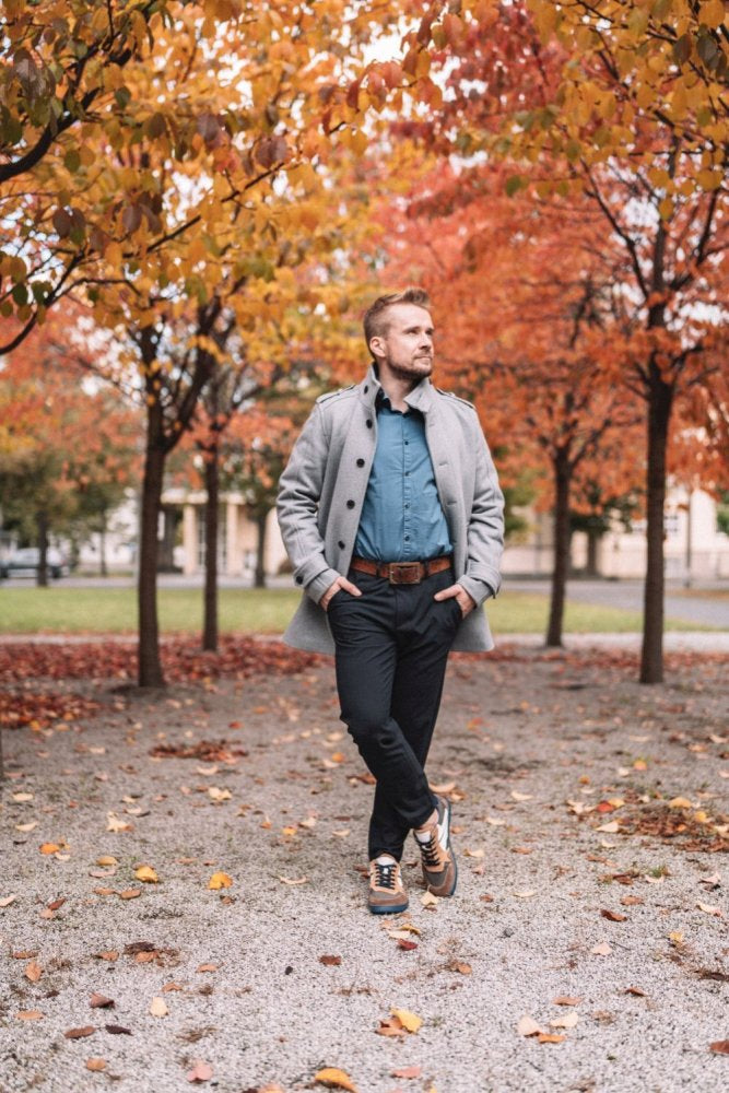 Eine Person in einem stylischen grauen Mantel, einem blauen Hemd und ZAQQ BREAQ Sierra-Sneakern steht auf einem Schotterweg, der von leuchtenden Herbstbäumen gesäumt ist. Der Boden ist mit Laub bedeckt, das durch das schlanke Design des Sneakers und die flexible ZAQQ-Sohle perfekt ergänzt wird und eine nahtlose Mischung aus Mode und herbstlicher Gelassenheit schafft.