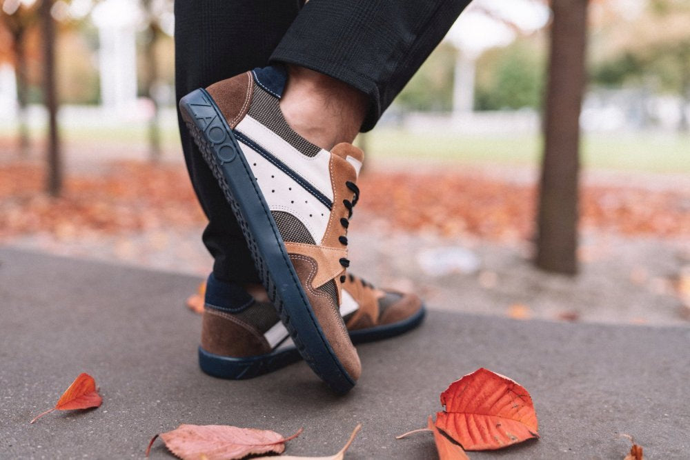 Eine Nahaufnahme zeigt eine Person, die die stylischen BREAQ Sierra-Sneaker von ZAQQ trägt, die eine Mischung aus Braun-, Schwarz- und Weißtönen aufweisen. Während sie auf einem mit leuchtend orangefarbenen und roten Herbstblättern übersäten Gehweg läuft, genießt sie dank der flexiblen Sohle des Schuhs den Komfort bei jedem Schritt. Im Hintergrund sind Bäume und Gras sanft verschwommen.