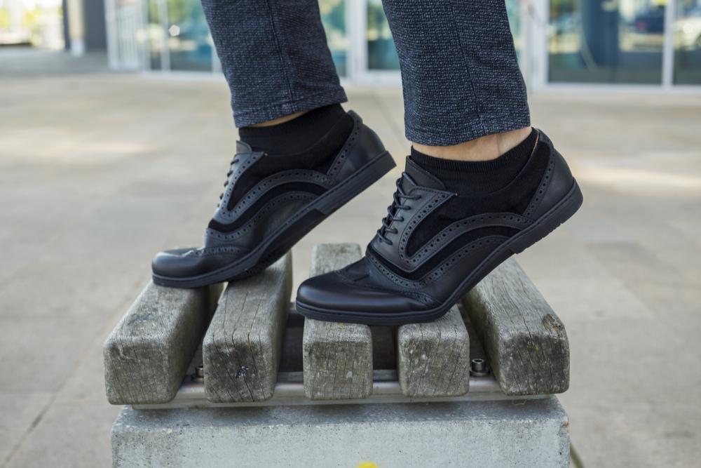 Eine Person in den ZAQQ BRIQ Brogue Deep Black Schuhen steht selbstbewusst auf einer Holzbank im Freien. Diese Schuhe sind mit dekorativen Perforationen versehen, die die dunkle, maßgeschneiderte Hose vor einem sanft verschwommenen Hintergrund perfekt ergänzen.