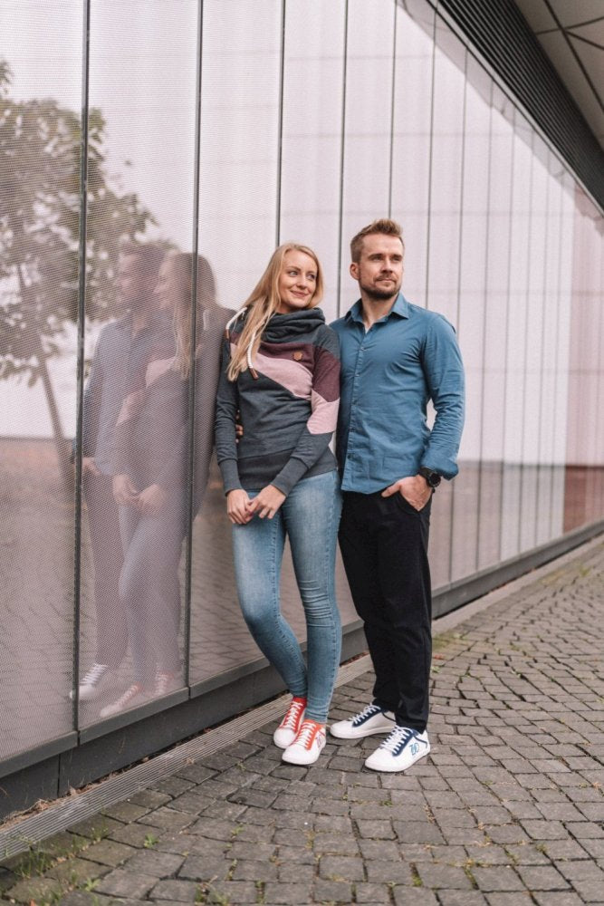 Ein Paar steht vor einem modernen Glasgebäude. Die Frau trägt einen grau-rosa Schal mit Jeans und bunten ZAQQ-Sneakers. Der Mann ist mit einem blauen Hemd, einer dunklen Hose und roten CEDRIQ-Sneakers von ZAQQ mit Kautschuksohlen bekleidet. Ihr Spiegelbild ist im Glas sichtbar.