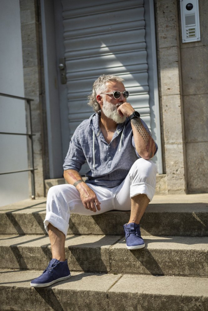 Ein bärtiger Mann mit Sonnenbrille sitzt draußen auf einer Treppe. Er trägt ein marineblaues Hemd von ZAQQ LANQASTER, weiße Hosen und blaue Schuhe. Er hat Tattoos und stützt sein Kinn auf seine Hand. Vor dem Hintergrund einer geschlossenen Metalltür wirkt er nachdenklich.