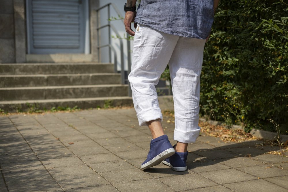 Eine Person in weißer Hose und ZAQQ LANQASTER Navy-Schuhen schlendert auf einem gepflasterten Weg, im Hintergrund führen Stufen zu einer schwebenden Metalltür. Grüne Sträucher zieren die rechte Seite und verstärken die ruhige Atmosphäre.