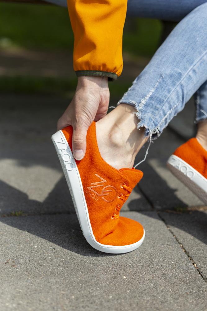 Eine Person in zerrissenen Jeans und orangefarbener Jacke sitzt auf einer Außenfläche und schlüpft in einen LIQE Orange Schuh von ZAQQ mit weißer Sohle. Der Schuh vermittelt ein Barfußgefühl, verfügt über ein sichtbares Logo und ist für eine optimale Temperaturregulierung konzipiert.