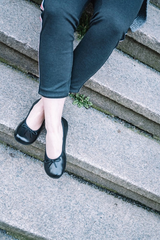 Eine Person sitzt auf einer Steintreppe und trägt schwarze LOOQ Flats aus weichem Nappaleder von ZAQQ und dunkle Leggings. Zwischen den Stufen wächst ein kleiner Abschnitt grünes Moos.