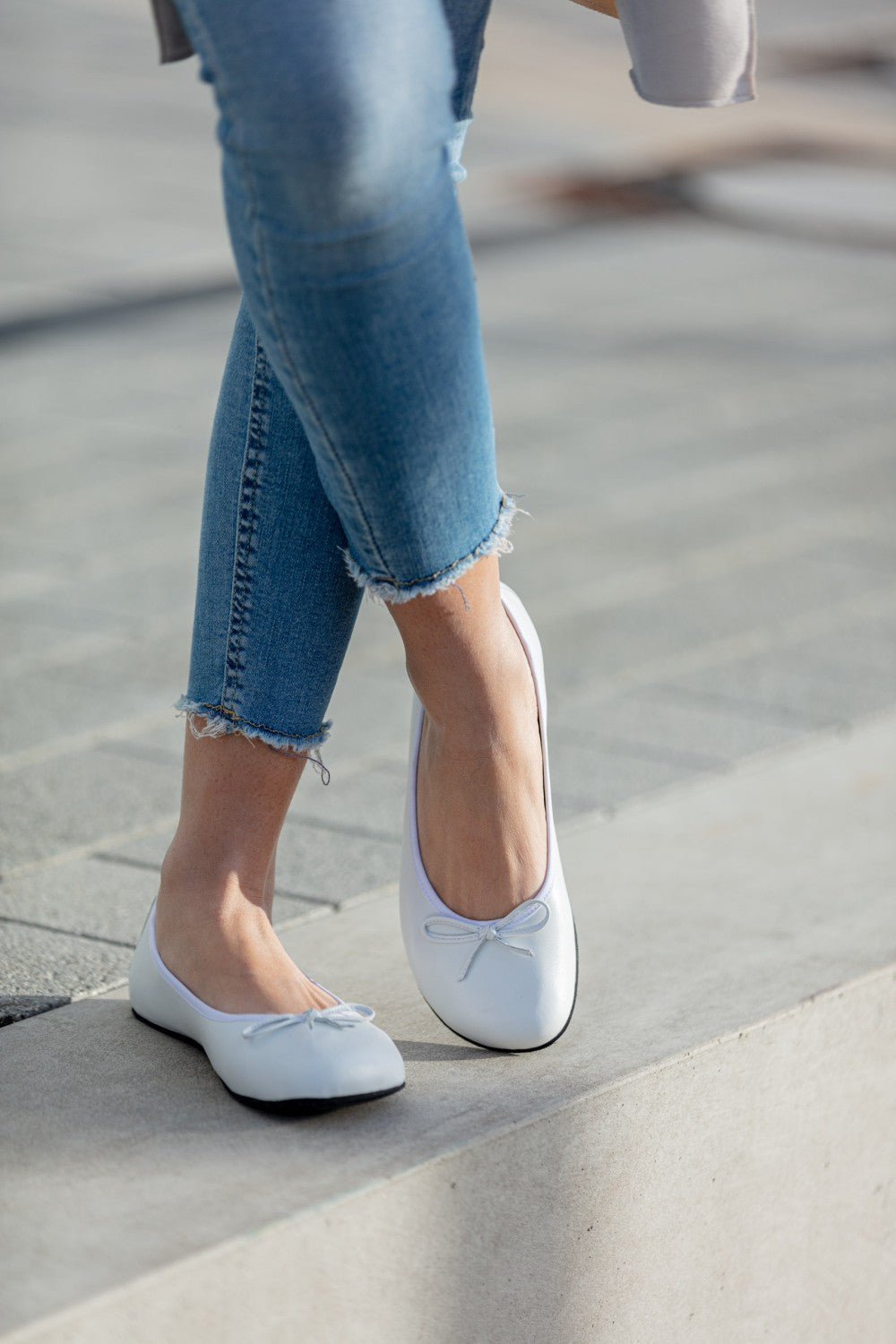 Eine Person in Bluejeans mit ausgefranstem Saum und weißen Ballerinas mit kleinen Schleifen von ZAQQ LOOQ läuft auf einem glatten Steinboden. Im Hintergrund ist der hellgraue Boden teilweise sichtbar.