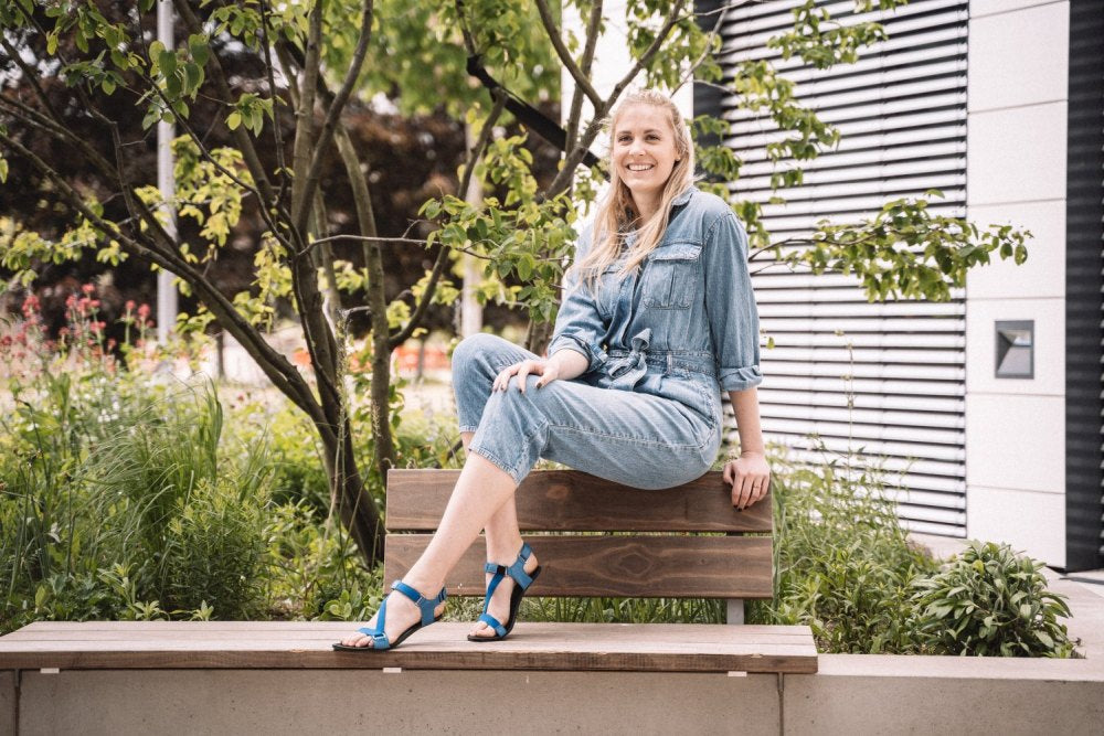 Eine Frau sitzt auf einer Gartenbank, bekleidet mit einem Jeansoverall und blauen MIQA Barfußsandalen mit strapazierfähiger Gummisohle von ZAQQ. Sie lächelt im Grünen, umgeben von Bäumen und Büschen.