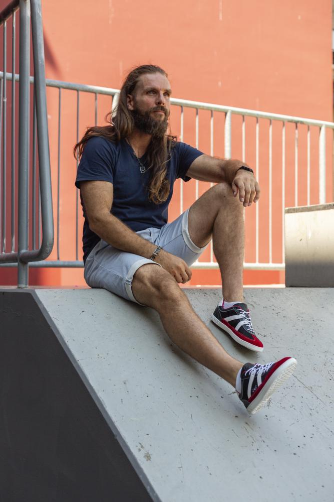 Ein Mann mit langen Haaren und Bart sitzt auf einer Betonkonstruktion. Er trägt ein marineblaues T-Shirt, hellgraue Shorts und rote OAQLEY-Sneaker von ZAQQ. Hinter ihm bildet ein Metallgeländer einen Kontrast zur rötlichen Wand. Die stylischen roten OAQLEY-Sneaker verleihen seinem lässigen Ensemble einen auffälligen Touch.