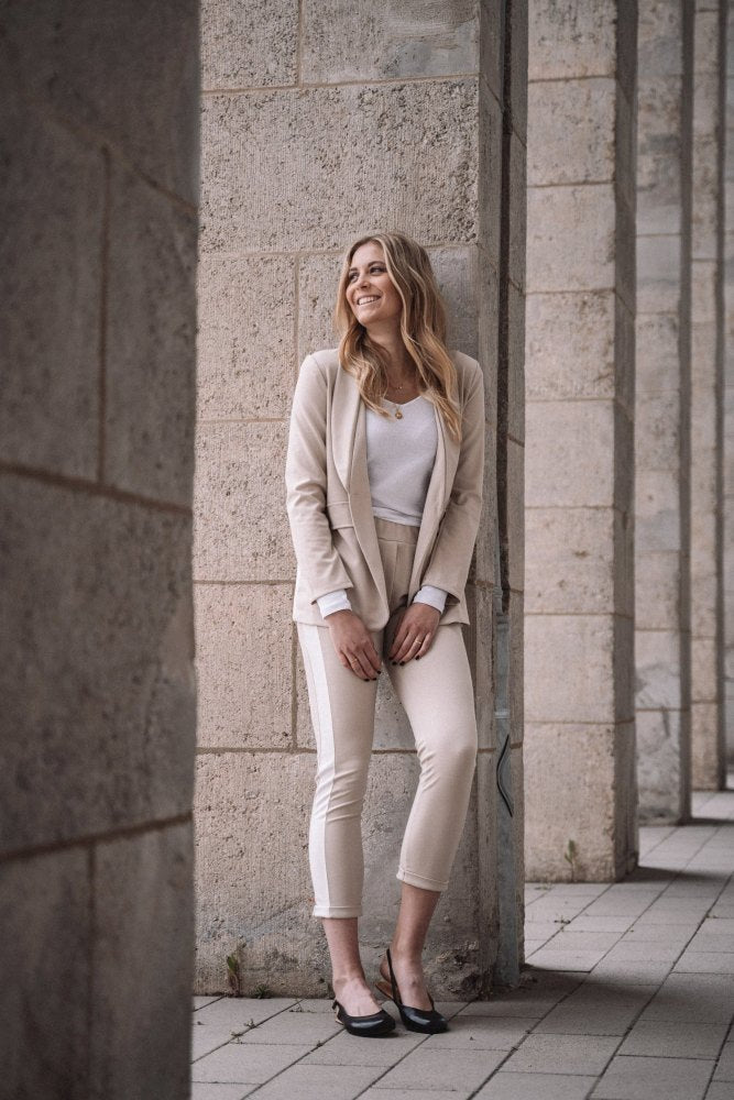 Eine Frau in einem hellen Anzug steht lächelnd vor einer Steinmauer, ihr Auftreten ist so anmutig wie das einer Ballerina. Sie steht draußen auf einem gepflasterten Platz mit hohen Säulen und fühlt sich entspannt und glücklich in ihren stylischen PINQ Black-Schuhen von ZAQQ.