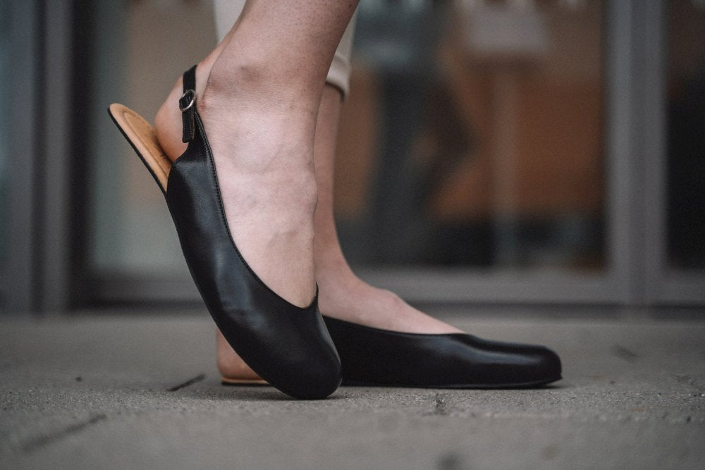 Eine Nahaufnahme einer Person, die die schwarzen Slingback-Schuhe ZAQQ PINQ trägt, die an die Anmut einer eleganten Ballerina erinnern und auf einer Betonfläche stehen. Der Hintergrund ist verschwommen und zeigt einen Teil eines Gebäudes mit glatten Glastüren.