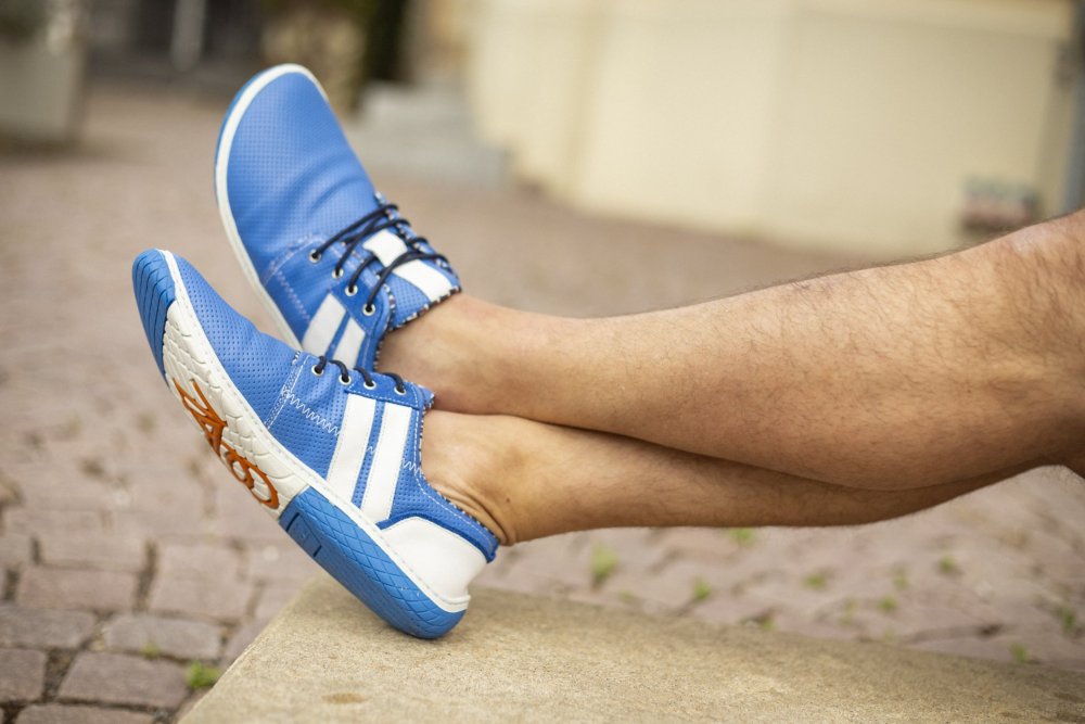 Eine Person sitzt lässig auf einer Steinbank und trägt ZAQQs QANBERRA Blue-Sneaker mit Seestern-Design auf der Sohle. Mit überkreuzten Beinen vor einem Hintergrund aus Kopfsteinpflaster und einem verschwommenen Gebäude verleiht der leuchtende Blauton ihrem entspannten Ensemble Flair.