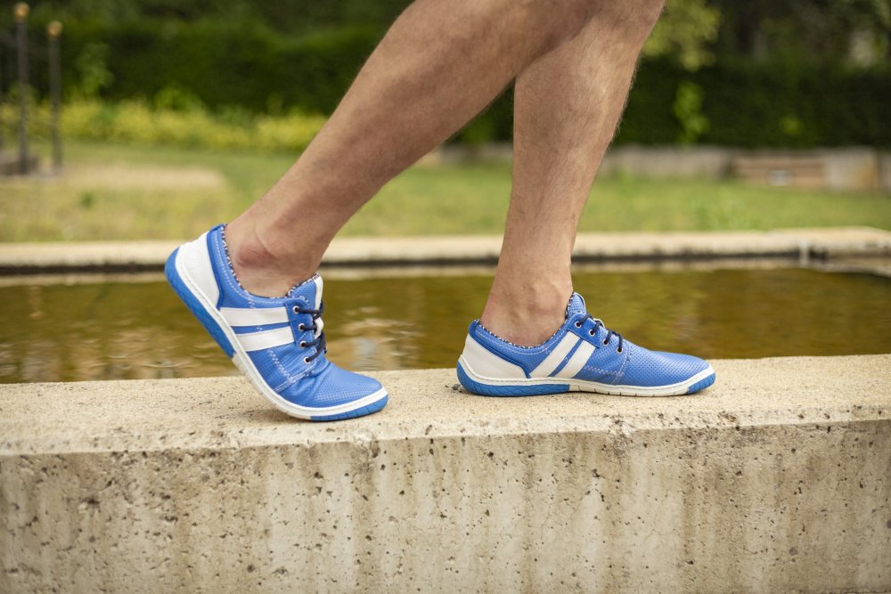 Eine Person schlendert auf einem Betonvorsprung neben einem Teich entlang und trägt die Sportschuhe QANBERRA Blue von ZAQQ. Das hochwertige Nappaleder-Design unterstreicht ihren Stil vor dem verschwommenen grünen Hintergrund.