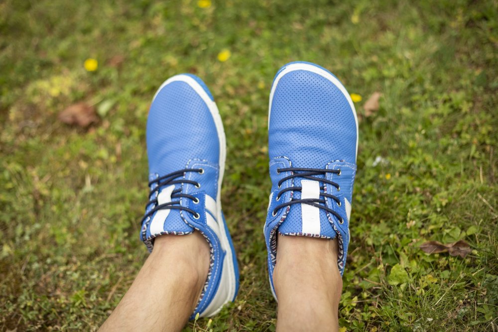 Eine Person, die die QANBERRA Blue-Sneakers von ZAQQ trägt, sitzt auf grünem Gras. Diese Schuhe haben schwarze Schnürsenkel und weiße Sohlen. Kleine gelbe Blumen und abgefallene Blätter sind verstreut und bilden einen reizvollen Kontrast zum üppigen Hintergrund.