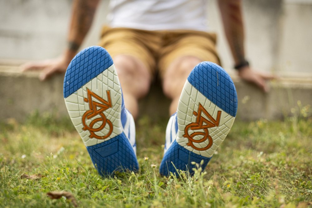 Eine Person sitzt auf einer niedrigen Betonmauer, die Beine sind auf dem Gras ausgestreckt, so dass die Sohlen ihrer Schuhe zu sehen sind. Diese ZAQQ QANBERRA Blue Barfußschuhe aus hochwertigem Nappaleder weisen ein blau-weißes Muster mit auffälligen orangefarbenen Buchstaben auf, die das Wort ZOO bilden. Die Person ist leger in Shorts gekleidet.