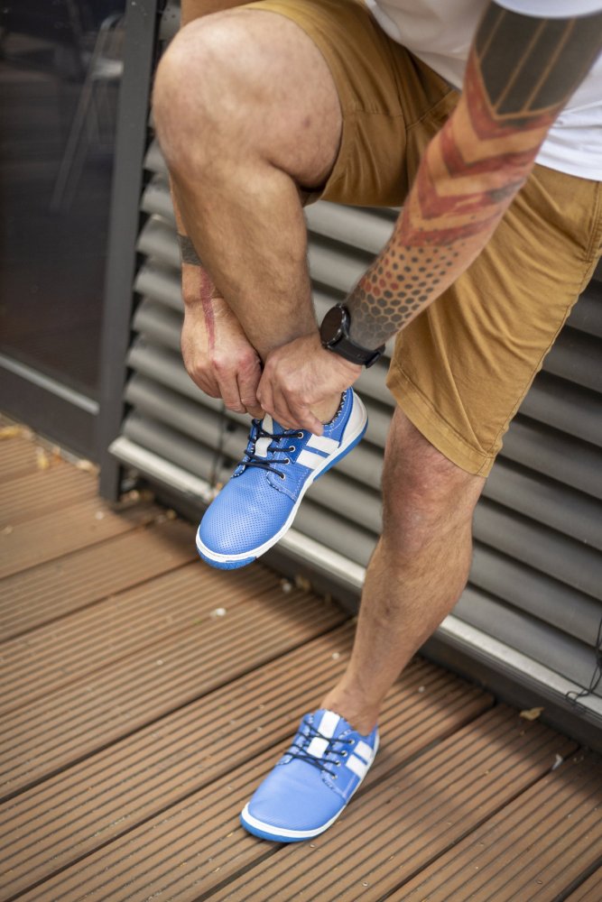 Eine Person mit tätowierten Armen und hellbraunen Shorts bindet die Schnürsenkel eines QANBERRA Blue-Sneakers von ZAQQ. Sie steht barfuß auf einem Holzdeck neben einer Metalllamellenwand und hebt das elegante Design und den Komfort hervor, der an Barfußschuhe erinnert.