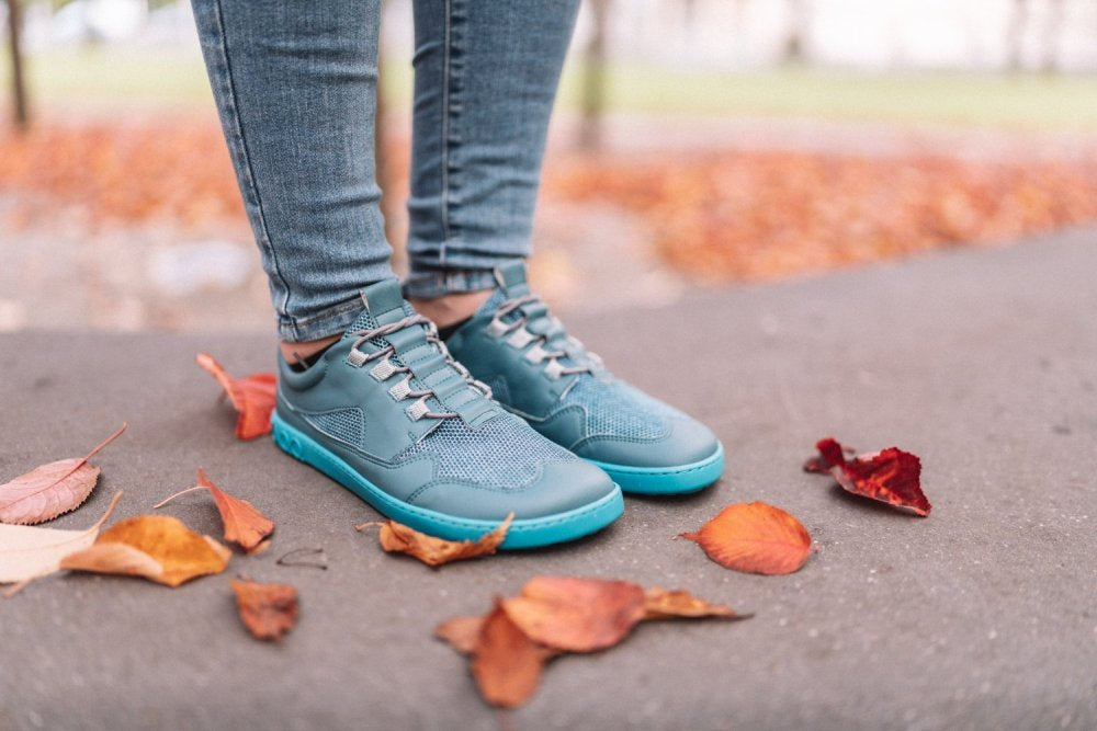 Eine Person in leuchtenden ZAQQ QARLY Turquoise Sneakers und Jeans steht auf einem Gehweg mit verstreuten Herbstblättern. Der Hintergrund ist unscharf, wodurch der stylische Barfuß-Sneaker von ZAQQ und das bunte Laub hervorgehoben werden.