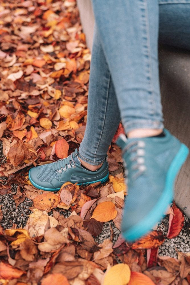 Eine Person, die die türkisfarbenen Sneakers „QARLY Turquoise“ und Jeans von ZAQQ trägt, sitzt auf einer Bank, umgeben von bunten Herbstblättern, die auf dem Boden verstreut sind.