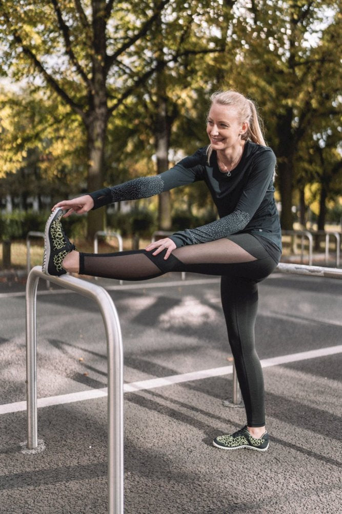 In der grünen Sportbekleidung QASEY von ZAQQ streckt eine Frau draußen auf einem Metallgeländer ihre Beine aus. Das Outfit aus atmungsaktivem Netzgewebe ergänzt ihr strahlendes Lächeln, während sie von Bäumen umgeben ist, durch deren Blätter das Sonnenlicht fällt. Die Szene scheint in einem Park oder auf einem Outdoor-Trainingsgelände angesiedelt zu sein.