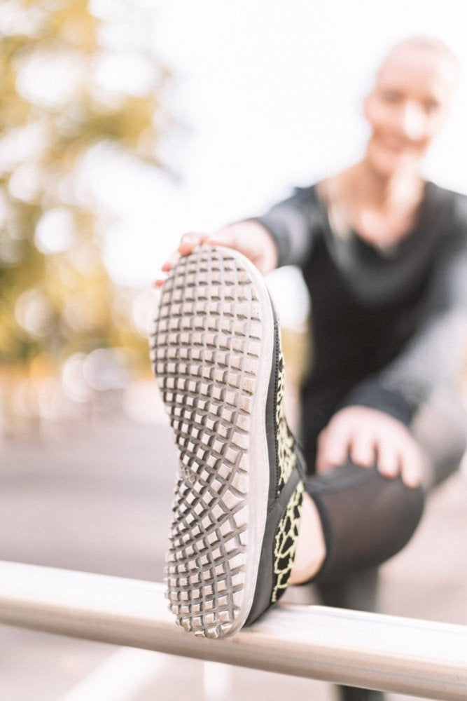 Eine Person in Trainingskleidung streckt ihr Bein auf einem Geländer aus und konzentriert sich dabei auf ihren ZAQQ QASEY Green Barfußschuh mit einem detaillierten Profilmuster aus veganen Materialien. Der Hintergrund ist sanft verschwommen und suggeriert eine Außenkulisse mit Bäumen.