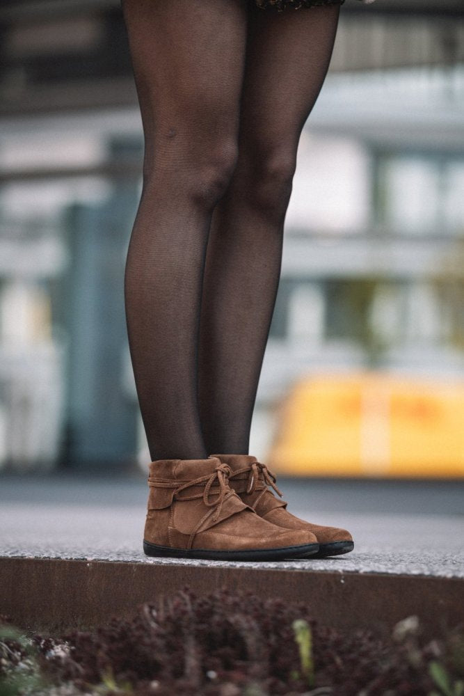 Eine Person in schwarzen Strumpfhosen und QEBEQ Cognac von ZAQQ steht auf einer Betonfläche. Der Hintergrund ist unscharf, um ein Gebäude mit großen Fenstern hervorzuheben.