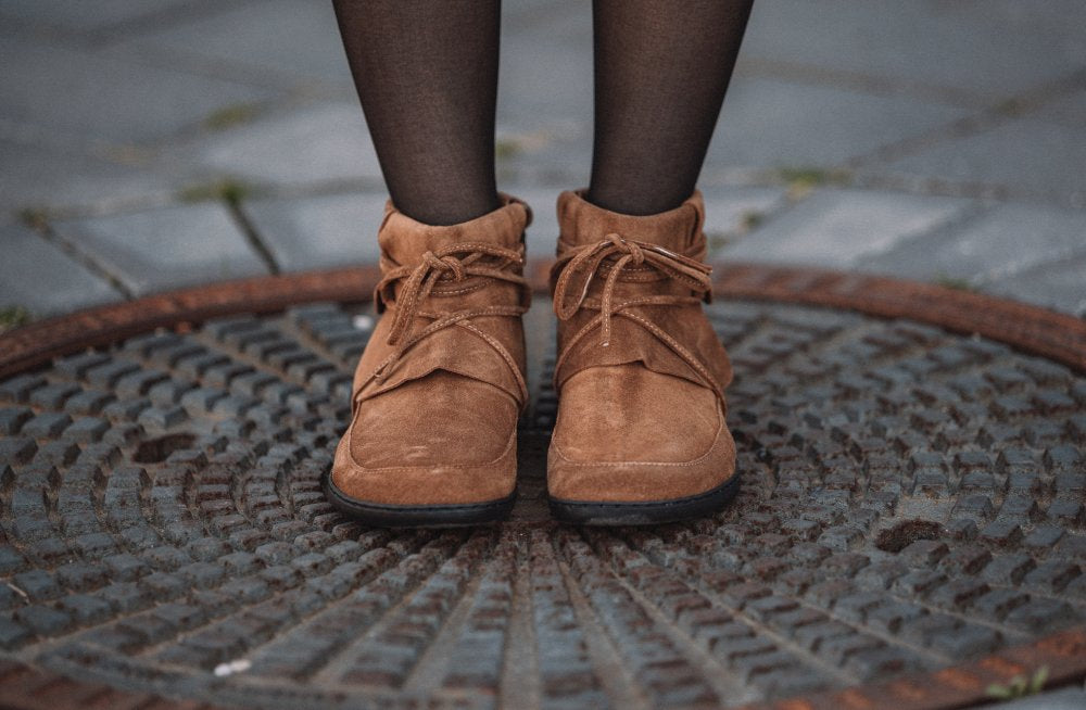 Eine Person in den Cognac-Wildlederstiefeletten QEBEQ von ZAQQ steht auf einem Metallgitter. Die Stiefel sind mit Schnürsenkeln geschnürt und die Person trägt dunkle Strumpfhosen. Der Boden ist mit grauen Steinen gepflastert.