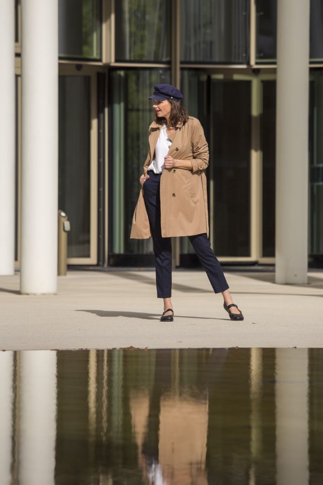 Eine Person in einem hellbraunen Trenchcoat, einer weißen Bluse, marineblauen Hosen und schwarzen DIEQO Nappa-Schuhen von ZAQQ steht selbstbewusst neben einem modernen Gebäude mit großen Fenstern, das sich in einem nahe gelegenen Gewässer spiegelt.