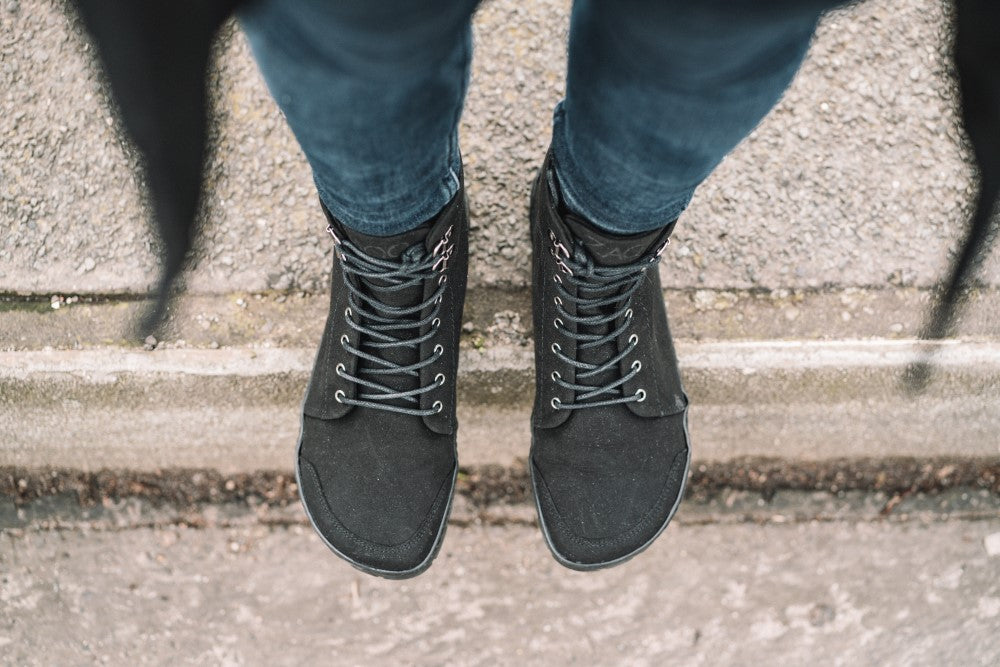 Luftaufnahme einer Person, die auf einem Betonbordstein steht. Sie trägt schwarze Schnürschuhe QICE von ZAQQ und blaue Jeans. Der Boden um den Bordstein herum ist strukturiert und rau.