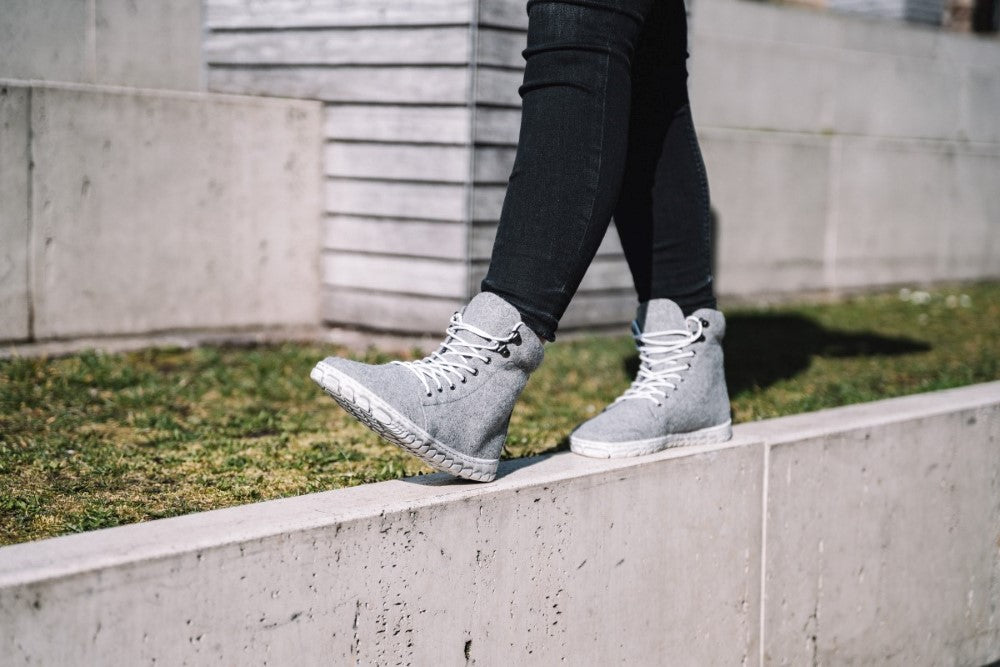 Eine Person trägt die stylischen ZAQQ QICE Grey High-Top Sneaker mit Outdoorsohle und schwarzer Hose und balanciert auf einer niedrigen Betonmauer. Im Hintergrund sind saftiges Gras und eine rustikale Holzkonstruktion zu sehen.