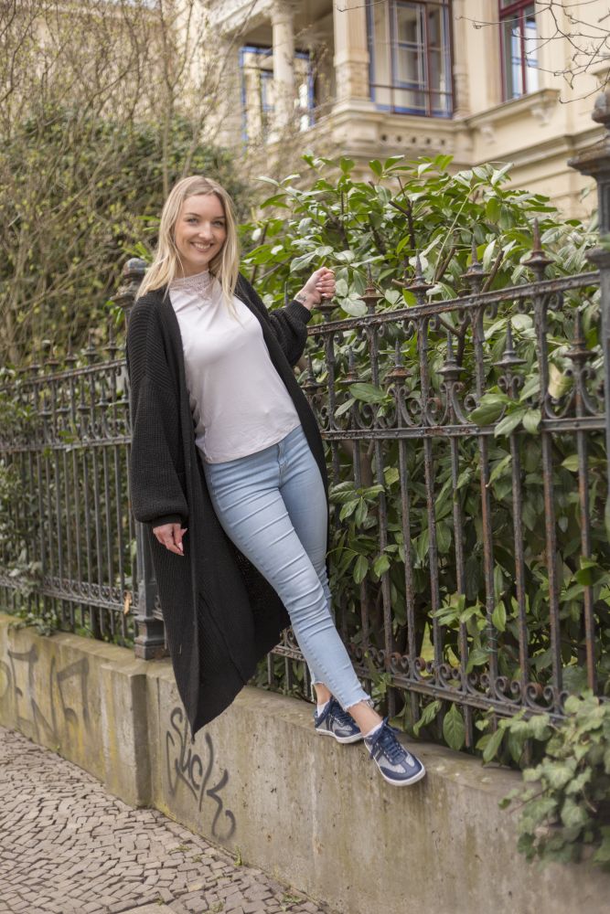 Eine Frau in einem schwarzen Mantel und ZAQQs QIMBERLY Blue Jeans lehnt an einem schmiedeeisernen Zaun. Sie lächelt und trägt stylische Barfuß-Sneakers, ein Fuß ruht auf dem Zaun. Im Hintergrund sind üppiges Grün und eine verzierte Gebäudefassade zu sehen.