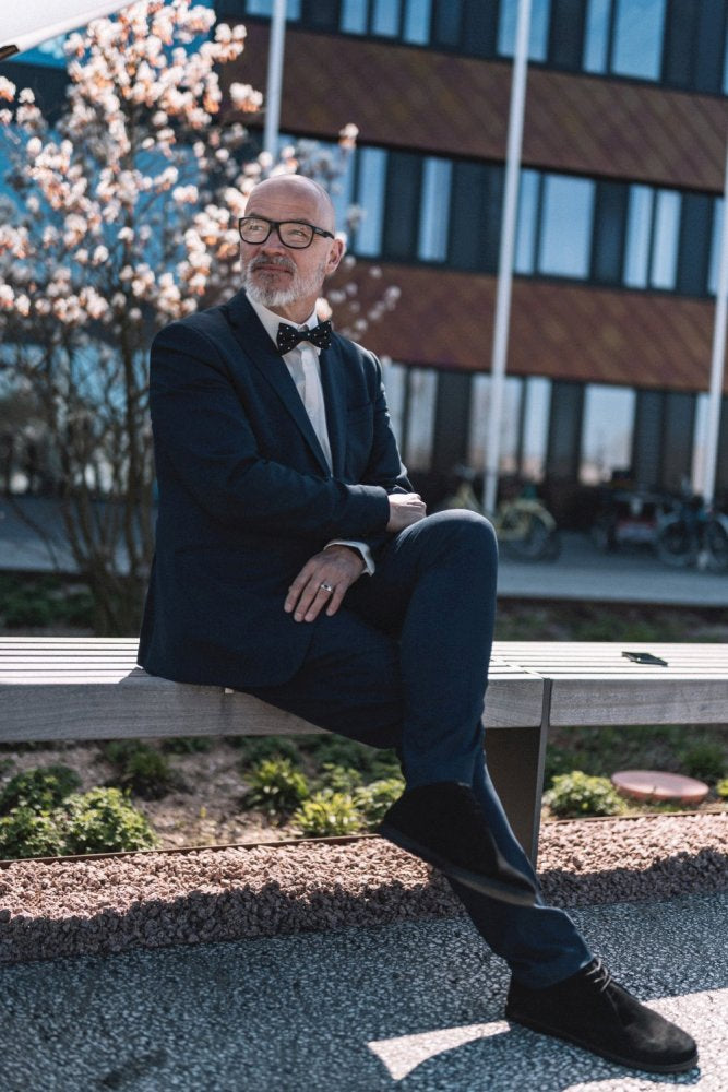 Ein Mann mit Brille und Bart, der einen Anzug und schwarze Schuhe von QLASSY der Marke ZAQQ trägt, sitzt auf einer Bank. Im Hintergrund sind ein modernes Gebäude und blühende Bäume zu sehen, was an eine Außenkulisse an einem sonnigen Tag erinnert.