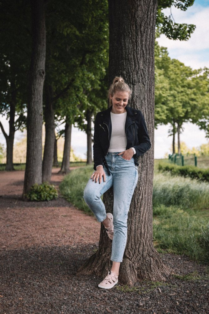 Eine lächelnde Person lehnt in einem Park an einem Baum. Sie trägt eine schwarze Jacke, ein weißes Hemd und hellblaue Jeans. Die QLEAR Light Pink Schuhe von ZAQQ bieten zusätzlichen Halt auf dem von weiteren Bäumen gesäumten Weg und sie genießt das saftig grüne Gras. Es ist ein sonniger Tag mit Wolken am Himmel.