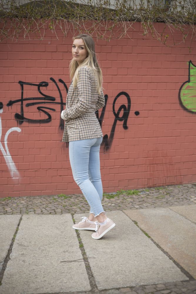 Eine Person mit langen Haaren posiert im Freien vor einer roten Backsteinmauer, die mit Graffiti bedeckt ist. Sie trägt einen karierten Blazer, hellblaue Jeans und QOMFY Wide Rosa-Schuhe von ZAQQ und blickt in die Kamera.