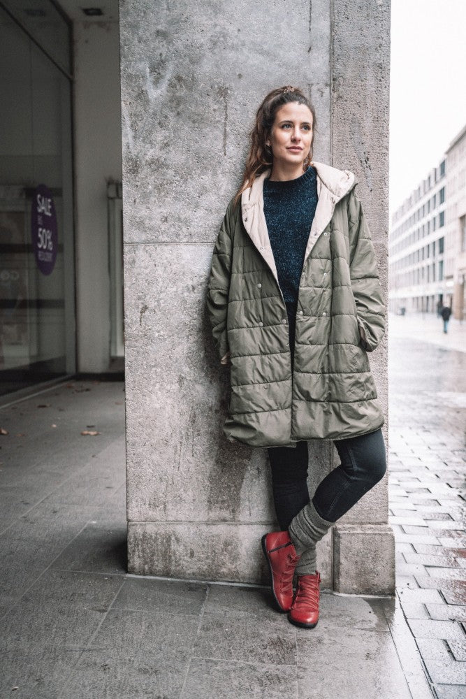 Eine Person mit langen Haaren lehnt an einer Betonwand. Sie trägt einen grünen Kapuzenmantel, schwarze Hosen, graue Beinwärmer und auffällige ZAQQ Quechee Velvet-Sneaker. Die Straße ist nass und im Hintergrund sind Gebäude zu sehen.