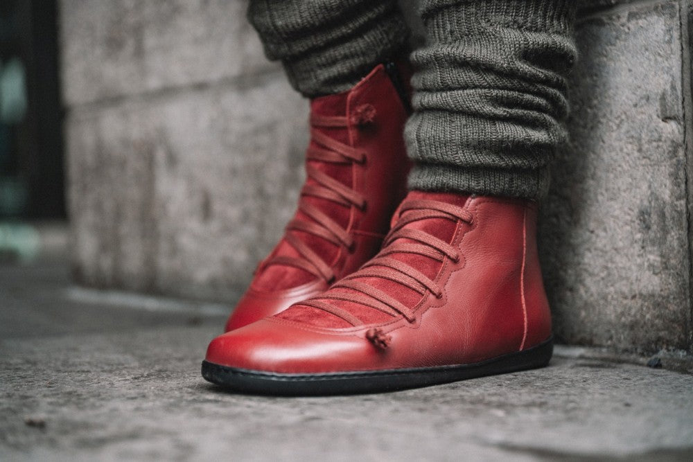 Nahaufnahme einer Person, die rote ZAQQ Quechee Velvet-Sneakers trägt, die Gebrauchsspuren aufweisen. Diese High-Top-Lederschuhe haben schwarze Sohlen und sind mit grauer, strukturierter Beinbekleidung kombiniert, während die Person auf einer Steinoberfläche vor einer Betonwand steht.