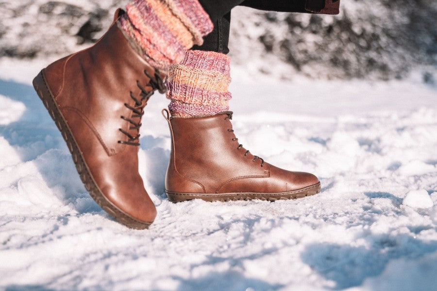 Ein Paar ZAQQ QUINTIC Winter Vegan Nut Stiefel, ergänzt durch bunte Stricksocken, macht sich auf den Weg durch eine schneebedeckte Landschaft mit malerischer Winterkulisse.