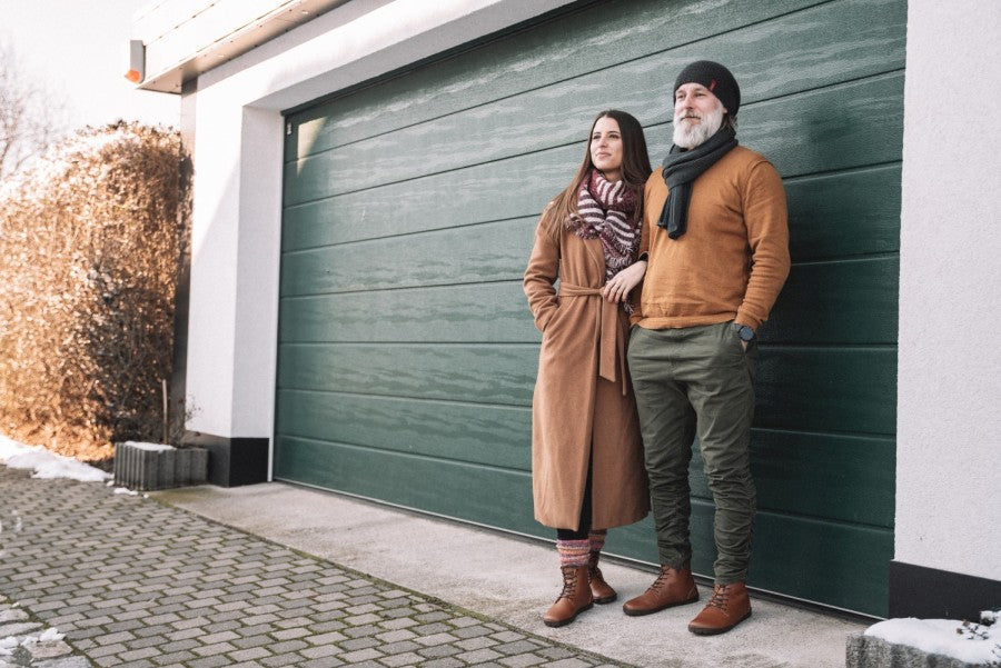 Ein Mann und eine Frau stehen vor einem grünen Garagentor. Sie trägt einen langen braunen Mantel, einen Schal und Stiefel mit Kautschuksohle. Er trägt einen braunen Pullover, eine grüne Hose und eine schwarze Beanie, wobei seine ZAQQ QUINTIC Winter Vegan Tabaco Barfußschuhe unter den Fransen des Schals hervorschauen. Beide wirken entspannt in der winterlichen Umgebung mit Schneeflecken auf dem Boden.