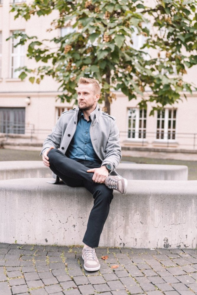 Eine Person mit kurzen Haaren und Bart sitzt mit übereinandergeschlagenen Beinen auf einer Betonbank. Sie trägt eine leichte Jacke, ein blaues Hemd, eine schwarze Hose und QUSQO Taupe Sneakers von ZAQQ aus atmungsaktivem Meshgewebe für zusätzlichen Komfort. Im Hintergrund sind ein großer Baum und ein Gebäude mit Fenstern zu sehen.