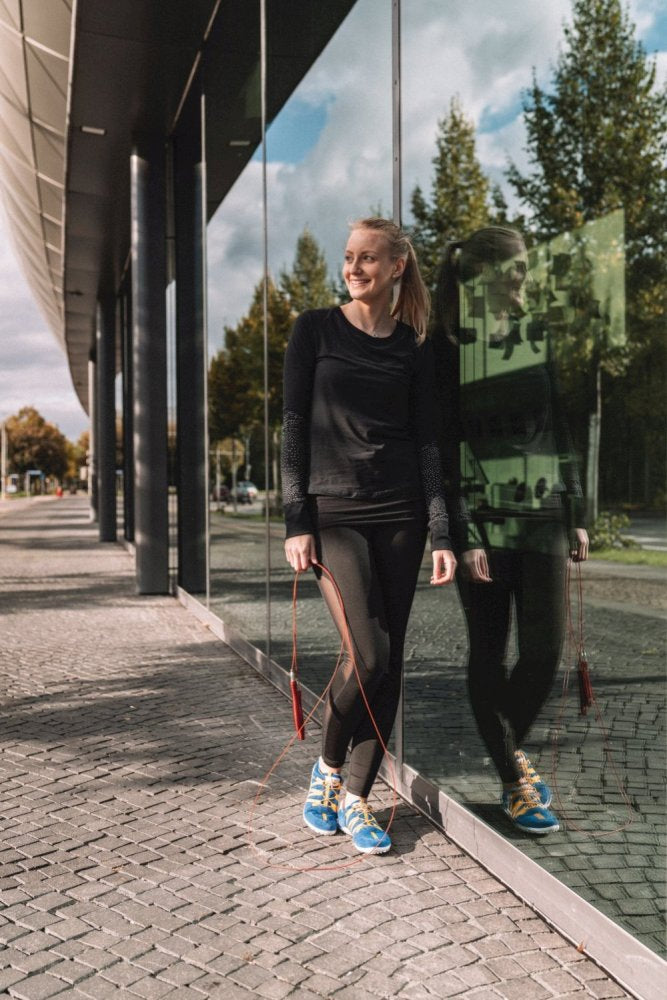 Eine Frau in schwarzer Sportkleidung steht mit einem Springseil lächelnd neben einem Glasgebäude. Ihr Spiegelbild ist im Glas sichtbar. Sie trägt blaue RIQ Blue Orange-Sneaker von ZAQQ und die Szene ist im Freien mit Bäumen im Hintergrund.