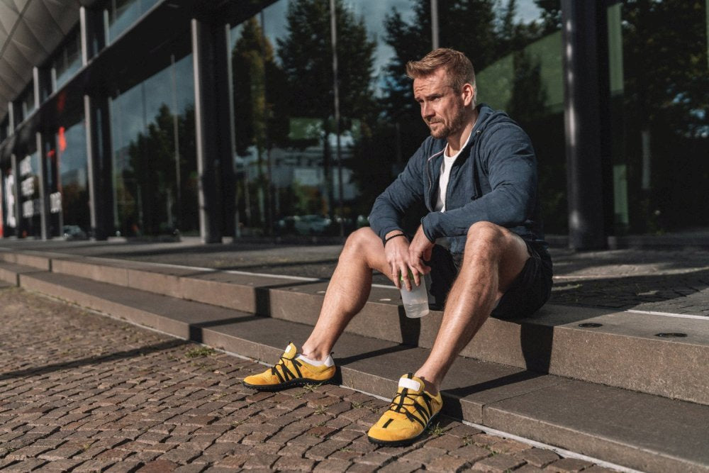 Ein Mann in Trainingskleidung sitzt auf den Stufen vor einem modernen Gebäude. Er trägt einen blauen Kapuzenpullover und Shorts und trägt die RIQ Yellow-Schuhe von ZAQQ für ein mühelos flexibles Tragegefühl. In seiner Hand ruht eine Wasserflasche, während sich in großen Glasfenstern die umliegenden Bäume spiegeln.