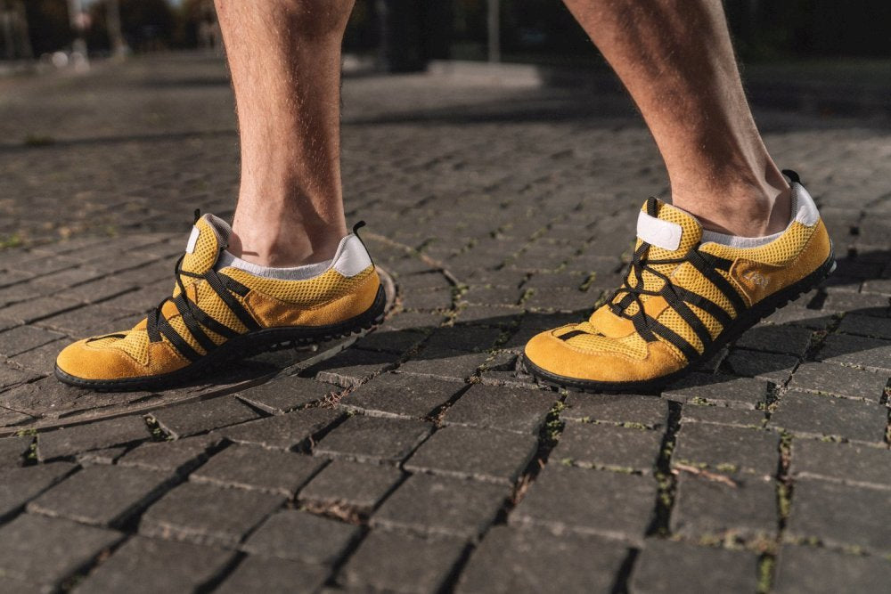 Eine Person, die die minimalistischen RIQ Yellow-Laufschuhe mit schwarzen Riemen von ZAQQ trägt, spaziert einen Kopfsteinpflasterweg entlang und hebt das Design und die Textur des Schuhs hervor. Diese Barfußschuhe bieten ein flexibles Tragegefühl und betonen die Füße und das Schuhwerk.