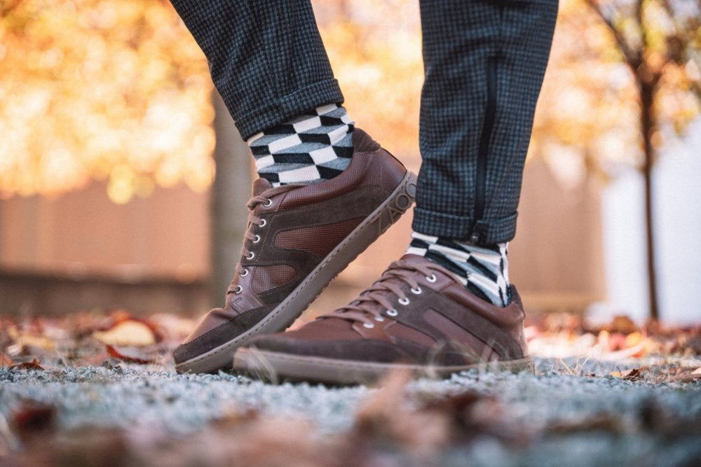 Nahaufnahme einer Person, die RIQO Brown-Sneaker von ZAQQ trägt, dazu schwarz-weiß karierte Socken und gemusterte Hosen. Der Hintergrund ist mit Herbstblättern und verschwommenen Bäumen geschmückt und ruft eine herbstliche Atmosphäre hervor, während die atmungsaktiven Materialien dieser Schuhe unvergleichlichen Komfort garantieren.