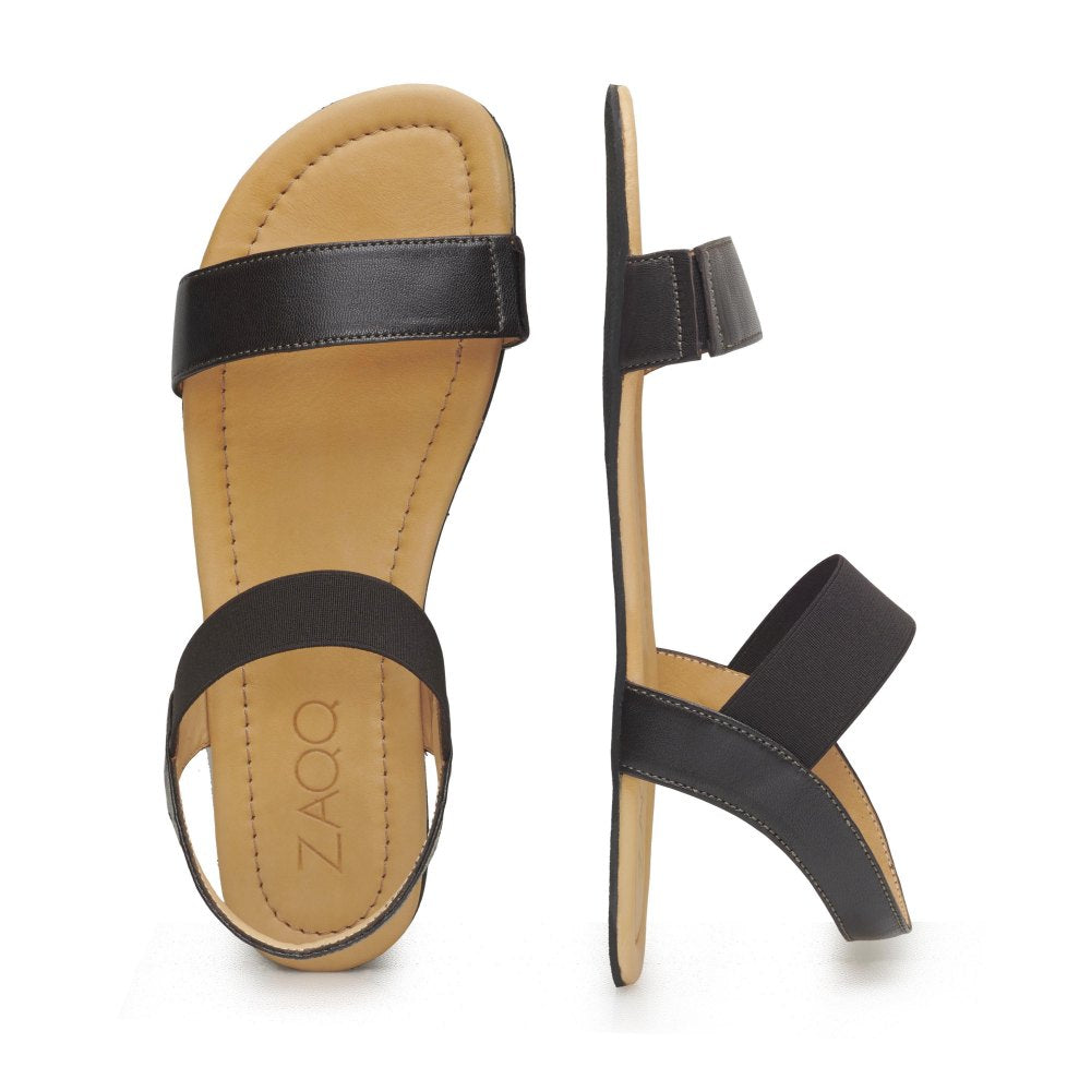 Gezeigt wird ein Paar eleganter dunkelbrauner Ledersandalen mit flacher Sohle. Eine Sandale ist von oben zu sehen, während die andere von der Seite präsentiert wird, was das minimalistische Riemendesign dieser dunkelbraunen SLIQ-Sandale hervorhebt. Der Markenname ZAQQ ist auf der Innensohle aus Nappaleder zu sehen.