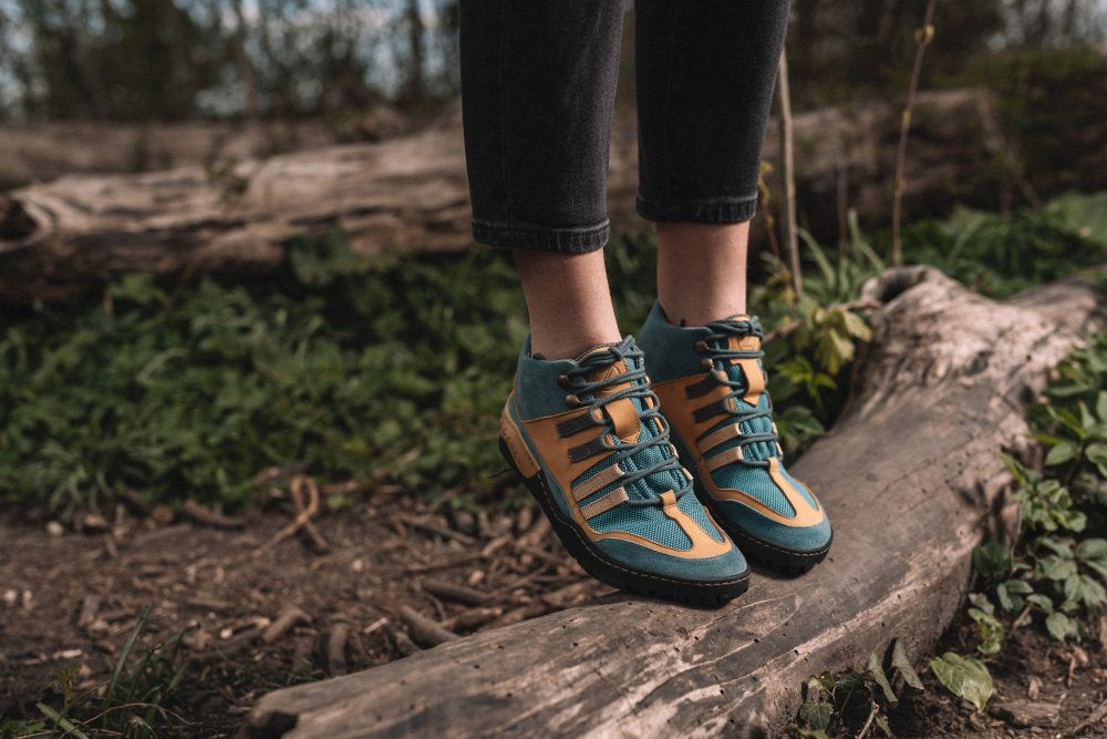 Eine Person in den wasserdichten Schuhen SQOUT Blue Orange von ZAQQ steht auf einem umgestürzten Baumstamm im Wald und fügt sich mühelos in das Grün ein. Die Schuhe haben schwarze Schnürsenkel und robuste Sohlen und bieten ein atmungsaktives Tragegefühl bei jedem Abenteuer.