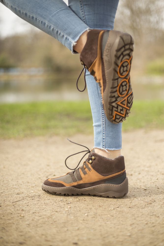 Eine Nahaufnahme zeigt eine Person, die auf einem Feldweg steht und die SQOUT Brown Waterproof Wanderschuhe von ZAQQ trägt. Die braunen und orangefarbenen Stiefel zeigen die ZAQQ TERRA Sohle mit sichtbarem Profil. Im Hintergrund schaffen verschwommenes Grün und Wasser eine ruhige Naturkulisse. Blue Jeans runden den Look ab.